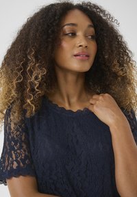 Top in pizzo blu navy con maniche corte e scallopate. Presenta intricati motivi floreali e un colletto delicato. Tessuto morbido e texture.