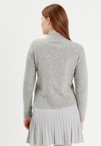 Pull en tricot gris avec col montant et ourlet côtelé, présentant une texture douce. Porté par-dessus une jupe plissée gris clair.
