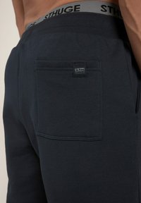 Dunkelblaue Jogginghosen bestehen aus einem weichen, strukturierten Stoff, haben einen elastischen Bund mit "STHUGE"-Branding und eine einzelne Gesäßtasche mit einem gestickten Etikett.