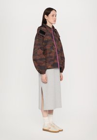 Braun camouflierter Jacke mit Kapuze, lila Reißverschluss und verstellbaren Manschetten, kombiniert mit einem grauen Midirock und beigen Sneakern.