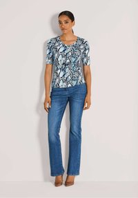 Kurzärmeliges Oberteil in Blau, Schwarz und weißem Schlangenprint, kombiniert mit dunkelblauen Schlagjeans und braunen High Heels; glattes Material und taillierter Schnitt.