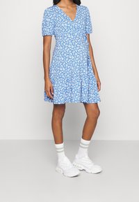 Donna che indossa un vestito avvolgente floreale azzurro chiaro con maniche a sbuffo, calze bianche con strisce grigie e sneakers bianche spesse su uno sfondo neutro.