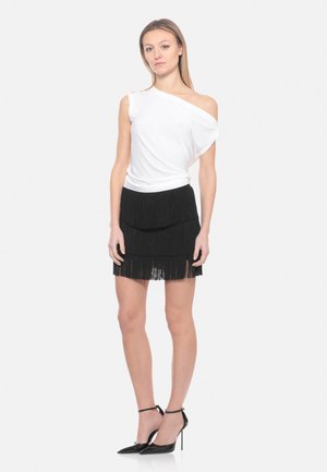 Donna che indossa un top bianco monospalla, minigonna nera con volant a strati e scarpe nere con tacco alto e cinturino alla caviglia, in piedi su sfondo bianco.