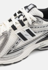Weißer und silberner Sportschuh mit einem Obermaterial aus Mesh, schwarzen Akzenten, gepolstertem Kragen und markantem Logo. Verfügt über eine strukturierte Sohle und Schnürsenkel.
