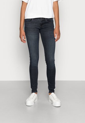 Personne debout portant un jean skinny foncé, des baskets blanches et un t-shirt blanc à manches courtes sur un fond clair uni.