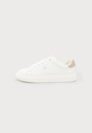Hvid lav sneaker med beige hælpatch, snørelukning, lille side logo og perforeringsdetaljer på en ensfarvet lys baggrund.