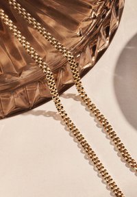 Eine goldene Kettenhalskette liegt über einer strukturierten, facettierten Glasschale. Die Kette hat einheitliche Glieder mit einem glänzenden Finish.