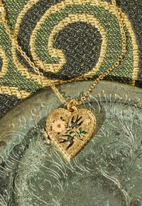 Zelta sirds kulons ar ziedu dizainu un divām melnām zvirbuļiem, kas ierakstīti teksturētā sudraba fonā.