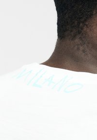 Primo piano del collo e della parte superiore della schiena di una persona che indossa una camicia bianca con la scritta "MILANO" in azzurro chiaro stampata vicino al colletto.