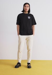 Black cotton t-shirt with a front graphic, paired with cream joggers. Model wears black shoes with a textured design, standing on a yellow surface.