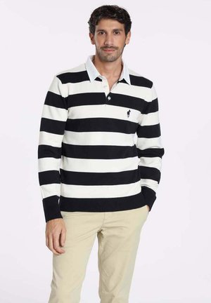 Pull-over à manches longues rayé noir et blanc avec un col, arborant un petit logo sur la poitrine et des poignets côtelés. Associé à un pantalon beige.