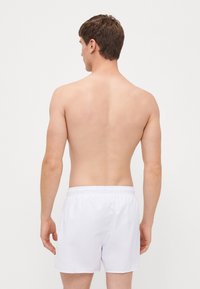 Fehér fürdőnadrág rugalmas derekú, könnyű anyagból készült, hátulról látható, meztelen bőrű férfi modellen.