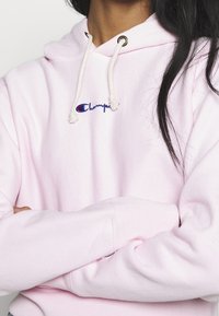 Sudadera de forro polar rosa con un bolsillo frontal, capucha con cordón y logo bordado en el pecho. Textura suave y corte relajado.