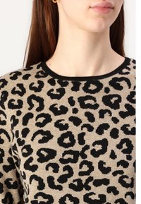 Kobieta nosząca beżowy sweter w czarne cętki i złoty kolczyk w kształcie koła, odsłaniając szyję i dolną część twarzy.