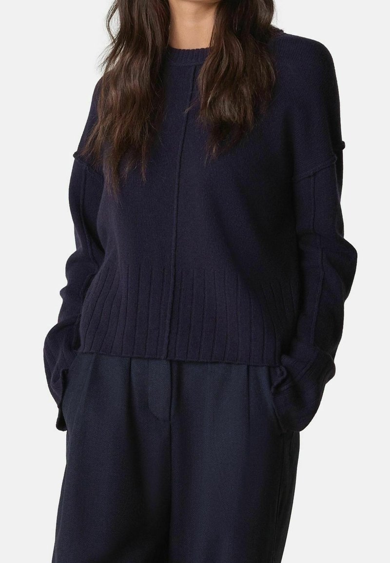 Femme portant un pull en maille côtelée bleu marine foncé à manches longues et un pantalon ample assorti, mains dans les poches, sur un fond uni.