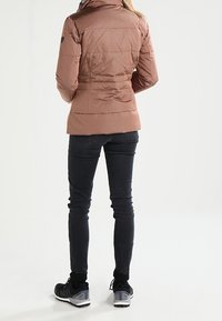 Chaqueta acolchada hasta la cadera en color marrón claro, con cuello alto y textura de canalé. Combinada con vaqueros negros pitillo y zapatillas deportivas.