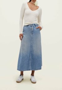 Vrouw in een wit geribbeld trui met lange mouwen, een lichtblauwe denim midi-rok, witte sneakers en een witte bloemenketting op een zwarte choker.
