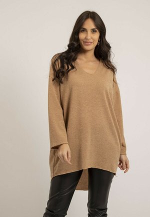 Femme aux longs cheveux foncés portant un pull ample à col en V beige et un pantalon en cuir noir, debout devant un fond clair.