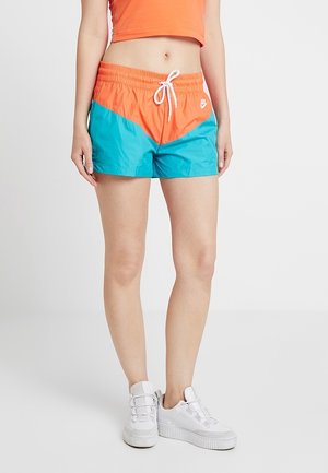 Femme portant un short Nike orange et turquoise avec un cordon blanc, des baskets blanches à plateforme, et un haut court orange.