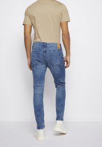 Jeans skinny de mezclilla azul con una cintura de media altura, que cuentan con bolsillos traseros y una etiqueta de marca de cuero marrón. Combinados con una camiseta beige y zapatillas blancas.
