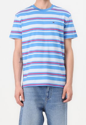 Homme portant un t-shirt à manches courtes rayé bleu, blanc et rouge, associé à un jean en denim bleu clair, debout devant un fond uni.