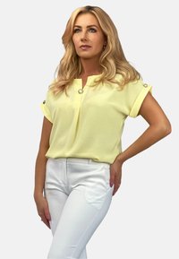 Żółta bluzka z krótkim rękawem, z dekoltem w serek i guzikami jako ozdobą. W połączeniu z białymi spodniami z bocznymi kieszeniami i dopasowanym krojem.