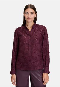 Donna che indossa una blusa button-up a maniche lunghe e texture, abbinata a pantaloni in pelle viola scuro, in piedi davanti a uno sfondo bianco.