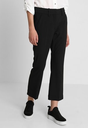 Personne portant un pantalon noir raccourci, une chemise blanche aux manches retroussées et des baskets noires à enfiler avec des semelles blanches.