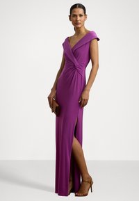 Vestido fúcsia com decote em V e detalhe torcido na cintura. Apresenta mangas curtas, uma fenda lateral e um design justo. Usado com saltos castanhos.