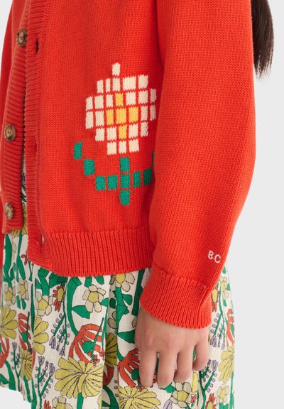 Cárdigan naranja tejido con patrón de flores pixeladas en blanco y amarillo, llevado sobre una falda con estampado floral; mano relajada al costado.