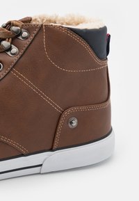 Brune lærhøyde sneaker med faux pelskrage, metalløyer og hvit gummisåle med svarte horisontale striper og sideakcent.