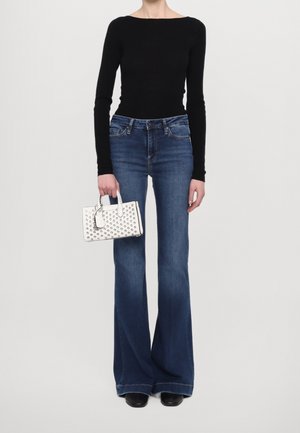 Sort langærmet bluse, blå flared jeans og en hvid håndtaske med hævet prikket mønster. Modellen står foran en ensfarvet baggrund.