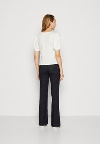 Blusa blanca texturizada con mangas abullonadas y detalles de volantes, combinada con jeans oscuros de campana y zapatillas blancas, vista desde la espalda.
