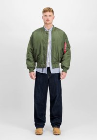 Bomber bunda olivově zelená se zipem vpředu, žebrovanými manžetami a červeným přívěskem. Natažena přes modrou pruhovanou košili a tmavé džíny.