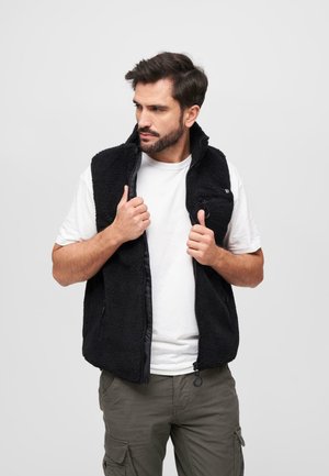Black fleece vest with a zip front, high collar, and a chest pocket; worn over a white t-shirt, featuring textured, soft fabric.