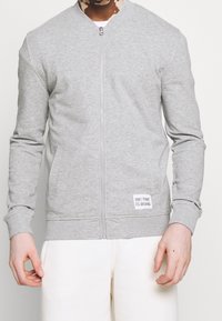 Persona che indossa una giacca grigia chiaro con zip e tasche anteriori, con un'etichetta che recita "NON ENTRARE NEL PANICO, È ORGANICO", abbinata a pantaloni bianchi.