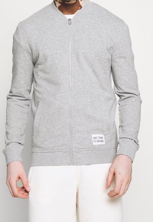 Person, der eine hellgraue Zip-Jacke mit Fronttaschen und einem Etikett mit der Aufschrift "KEINE PANIK, ES IST BIO" trägt, kombiniert mit weißen Hosen.