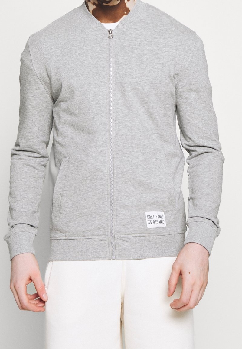 Persona che indossa una giacca grigia chiaro con zip e tasche anteriori, con un'etichetta che recita "NON ENTRARE NEL PANICO, È ORGANICO", abbinata a pantaloni bianchi.