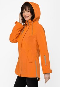 Oranssi pehmeäkuorijakku hupulla, jossa on vetoketjulliset taskut, harmaat yksityiskohdat ja istuva muotoilu. Sileä pinta, kevyttä materiaalia.