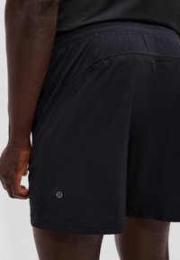 Black athletic shorts with a small reflective logo on the left thigh, worn by a person with dark skin, shown from the back waist to mid-thigh.