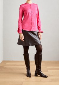 Mujer vestida con una blusa rosa de manga larga, falda de cuero negra, medias negras y botas altas de cuero negro, de pie sobre un suelo de madera.