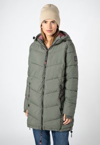 Vrouw in een groene gewatteerde jas met capuchon en rits aan de voorkant, met een beige gebreide muts, staand tegen een effen lichte achtergrond.