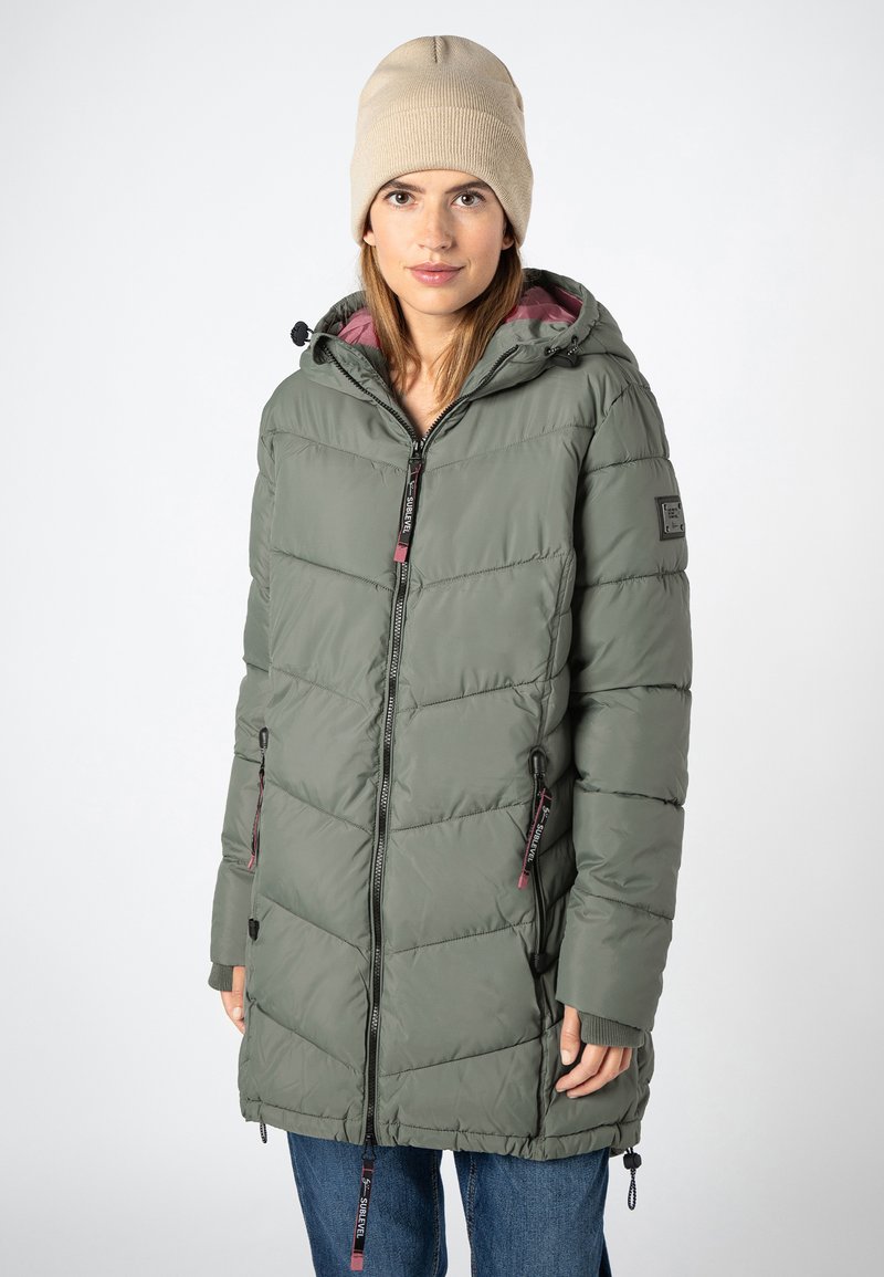 Frau in einer grünen, gefütterten Jacke mit Kapuze und Frontreißverschluss, die gegen einen schlicht hellen Hintergrund steht, mit einer beigen Strickmütze.