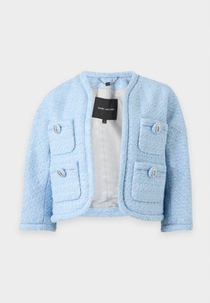 Femme portant un tailleur jupe bleu clair texturé avec quatre poches avant et une chemise blanche, debout avec des chaussures noires à bout pointu sur fond blanc.