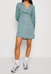 Pitkähihainen kietaisumekko sinisessä ja vihreässä geometrisessä kuosissa, V-pääntiellä ja levenevällä hameella. Yhdistetty urheilullisiin harmaisiin lenkkitossuihin.