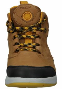 Scarpa mid-top in pelle marrone con lacci a scacchi neri e gialli, un logo tondo sulla linguetta e una suola a contrasto nera e bianca.
