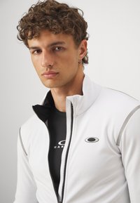 Hvit zip-up jakke med grå detaljer, laget av glatt materiale, lagdelt over en svart topp med et logo. Nærbilde.