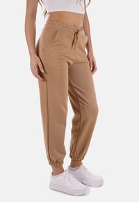 Pantaloni jogger marroni con vita elastica e caviglie a costina, dotati di tasche laterali e cordoncino in vita. Abbinati a sneakers bianche.