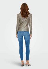 Metallic langesloefhemd met verticale strepen; nauwsluitend ontwerp. Draagt bij blauwe skinny jeans en zilveren hoge haksandalen, gezien vanuit de achterkant.