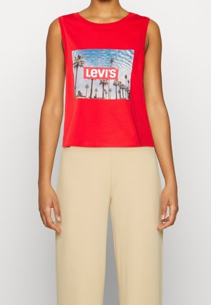 Femme portant un haut sans manches rouge Levi's avec un motif de palmiers et ciel bleu, associé à un pantalon beige taille haute.